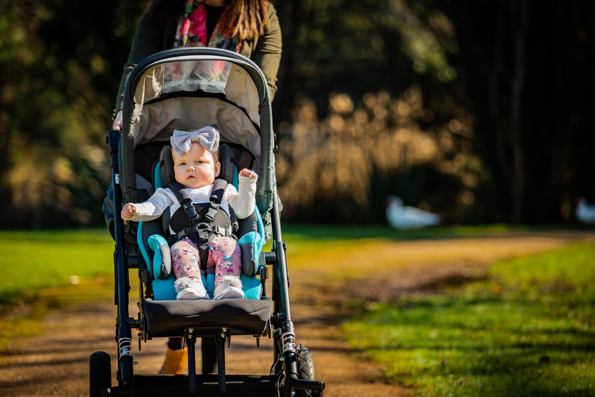 Lady pushing toddler in a black mini stroller with teal blue seat cushion and back rest and sun hood at a park.