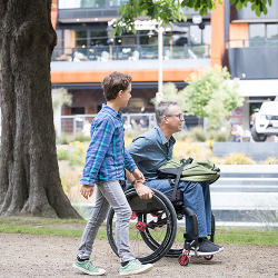 Young boy walking beside middle-aged man in a manual wheel car with an item on his lap help in place with a lap stacker strap.