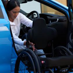 Lady sitting in a blue car with door open and Zygo wheel chair outside with her holding on to the back of it.