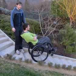 Man sitting in manual wheelchair with his arms crossed balancing on his two back wheels on stairs with another man standing beside him.