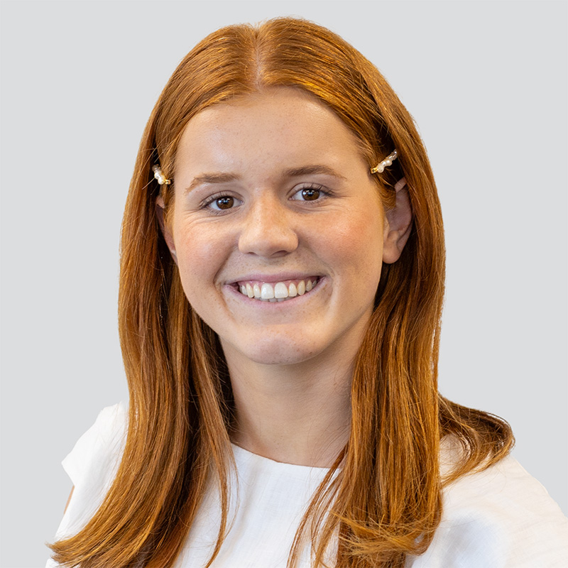Headshot of young woman named Ashleigh Scarlett, smiling into camera. Ginger hair, brown eyes, white blouse, grey background.