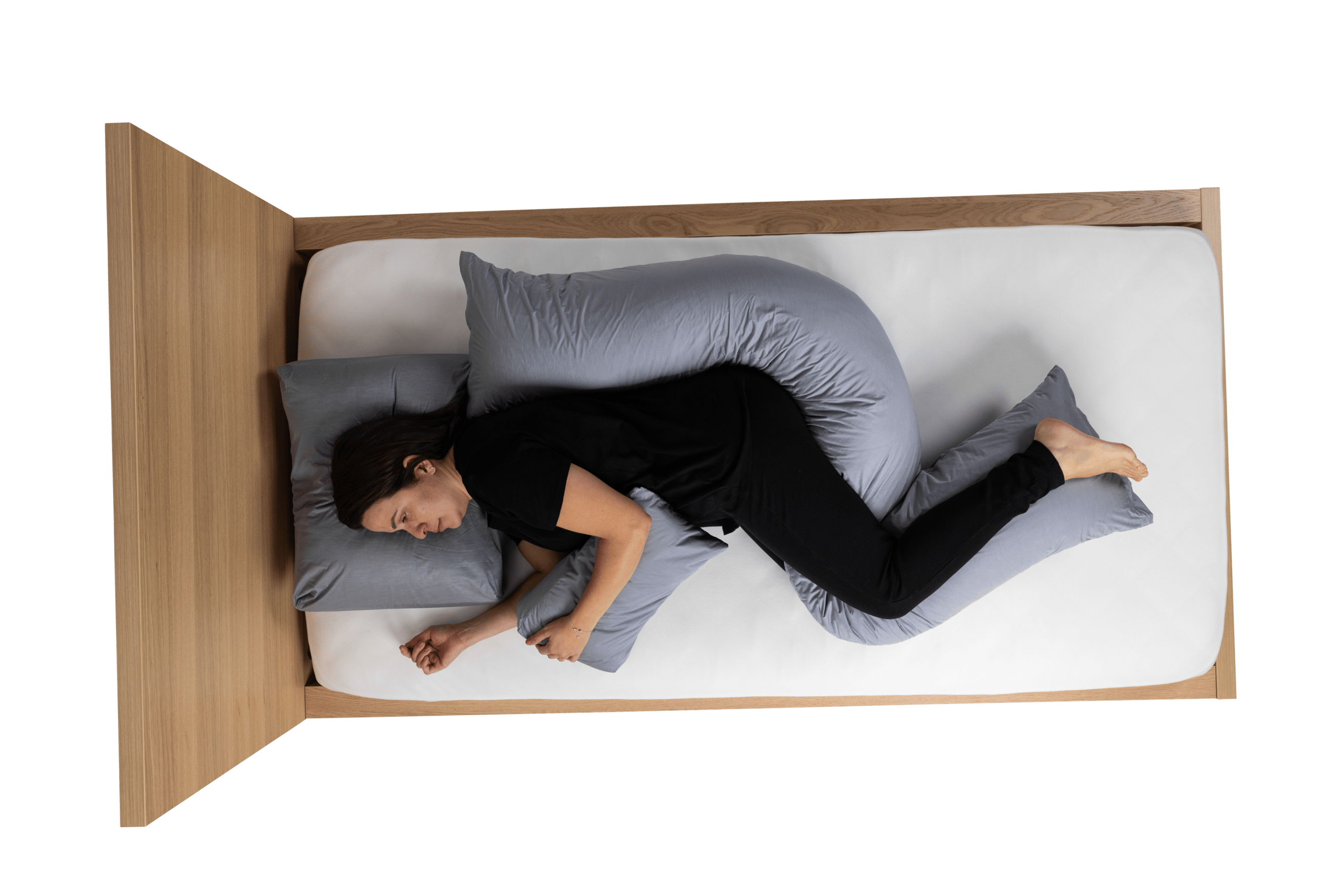 Aerial view of a woman in black clothing lying on a wooden bed with a white mattress, supported by grey Poz'In'Form postural support cushions - specifically an occipital cushion under her head, a hand wedge cushion supporting her left hand, and a decubitus cushion supporting her back and legs.
