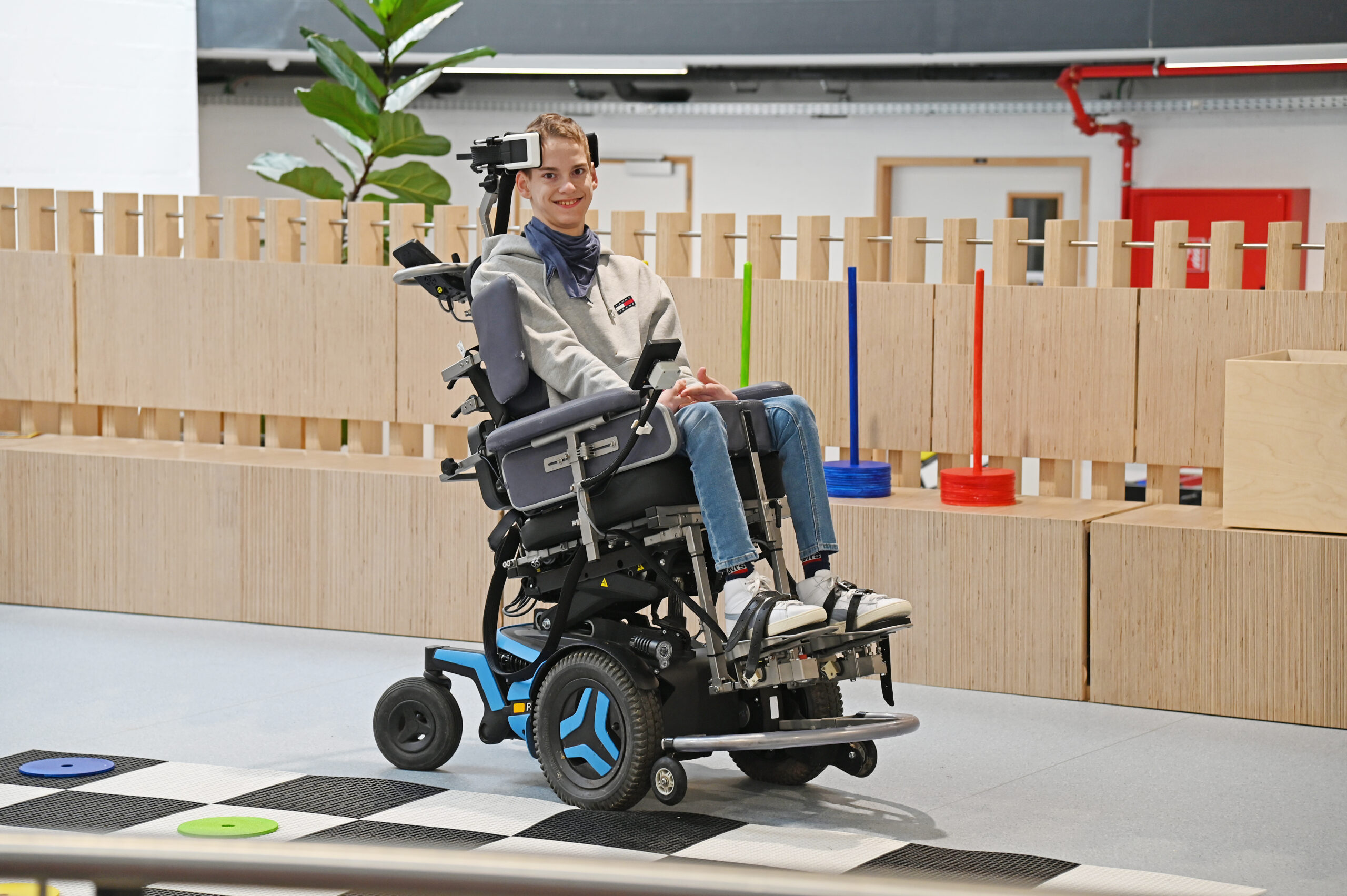 Full length image of a boy in a power wheelchair, smiling at the camera as he uses his CoMoveIT head control to move the wheelchair towards the camera.